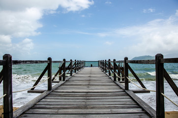 Old wooden pier by the sea. A pier with wooden railing, a rope is stretched along the railing. White waves on the seashore. On the horizon is a blue sky with white clouds. Perspective. A place of