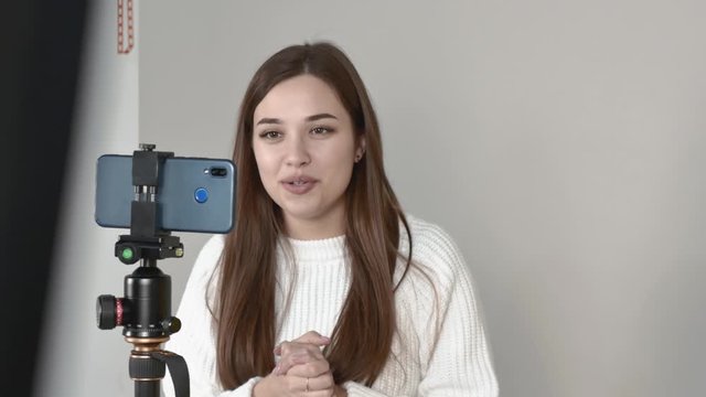 Grey background Caucasian girl blogger white jumper Straight long hair brown haired, Standing in front of phone camera, talking, laughing. The left corner is blocked by a black lighting fixture
