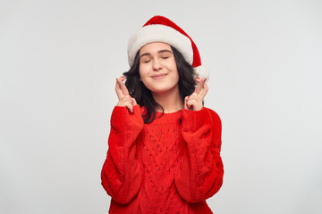 Girl in a red sweater and Santa Claus hat holding fingers crossed dreaming about the best gift