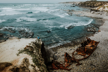 Fototapeta na wymiar Woman sitting next to shipwreck on the coast in Portugal