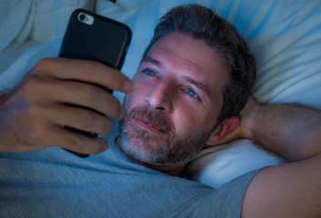 Obraz premium happy man with blue eyes lying on bed late at night in dark light networking on mobile phone or online dating smiling relaxed enjoying internet in social media addiction