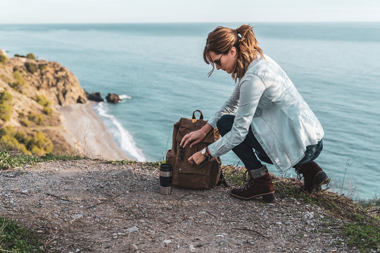 Young hip woman looking in her backpack explores the coast on a beautiful day. Concept of exploration and adventures