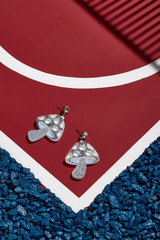 Object shot of pop-art earrings in a shape of silver glittering mushroom, lying on a red surface with white stripes and blue stones on borders. The upper part of earrings is a shiny rhinestone.
