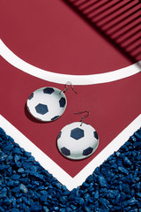 Object shot of leather pop-art football ball earrings, lying on a red surface with white stripes and blue stones on borders. 