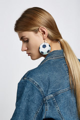 Profile portrait of a blond girl, wearing jean jacket. She has leather pop-art football ball earrings. Girl is looking down.