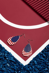 Object shot of pop-art earrings with double leather tearshaped pendant with American flag print, lying on a red surface with white stripes and blue stones on borders. 