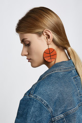 Profile portrait of a blond girl, wearing jean jacket. She has white leather pop-art basketball ball earrings. Girl is looking down.