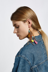 Profile portrait of a blond girl, wearing jean jacket. She has pop-art earring decorating with leather stripes, beads, fabric elements and chains. Girl is looking down.