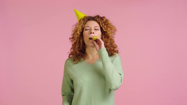 happy woman waving hand and blowing in party horn isolated on pink