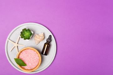 Beauty and spa concept. Sea salt, oil, green leaves and shells on grey tray on purple background. Flat lay. Copy space. 