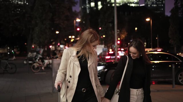 Two attractive girls are rolling their suitcases in night city. Females hold hands, smiling, enjoying walk together, wearing fashionable clothes.