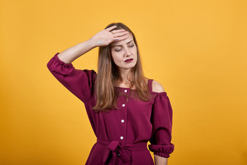 Woman with tired and sick expression holds der hand on forehead. She is tired and maybe has fever. She dressed up in burgundy bluse. Behind her there is orange background.