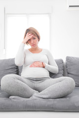 Fototapeta premium Pregnant tired exhausted woman with stomach and head issues at home on a couch, being sick.