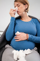 Fototapeta premium Pregnant woman catching cold, flu, virus, sitting at home on a couch, blowing nose and beaing sick.