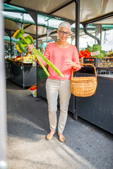 Senior woman buying fresh organic produce and flowers at a local farmer's market.  Senior woman holding basket full of groceries and flowers, in nature on sunny summer day