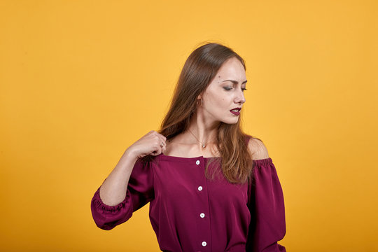 Ginger girl in burgundy bluse with bow over isolated orange background thoughtfully looks right holding her fist on shoulder