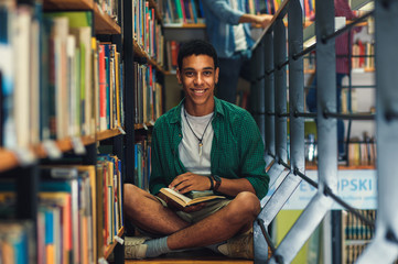 Young male student study in the library reading book while sitting near bookshelf.