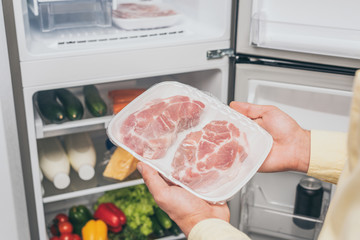 cropped view of man holding frozen meat near open fridge full of food
