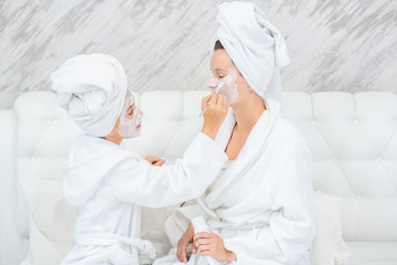 Young girl puts white cream on mother`s face at home. Mom and child girl are in bathrobes and with towels on their heads