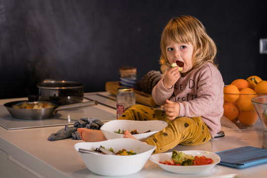 Little girl sitting on top of the kitchen surrounded by vegetable dishes and eating a piece of apple