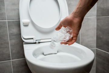 cropped view of man throwing crumpled plastic bottle in toilet bowl in modern restroom with grey tile