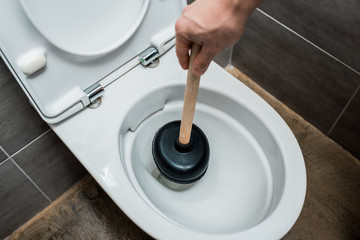 cropped view of plumber using plunger in toilet bowl in modern restroom with grey tile