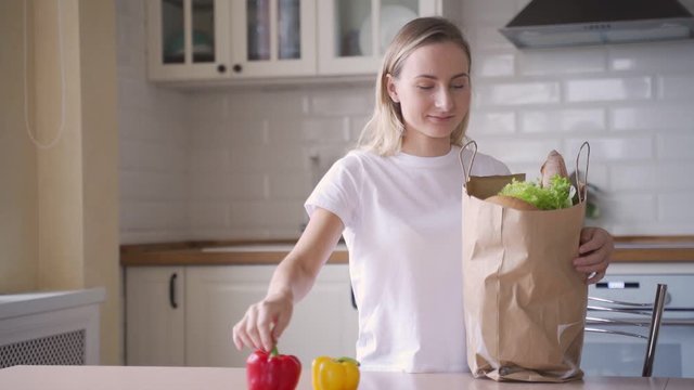 Happy to unwind the young woman holding groceries paper bag in the kitchen. A woman unpacks vegetables and fruits from a supermarket bag.