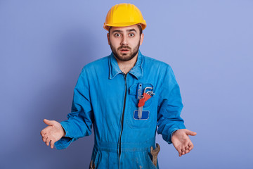 Astonished shocked young builder wearing yellow helmet and blue uniform, standing with opened mouth and spreading hands, making helpless gesture, handsome guy looks surprised. People emotions concept.