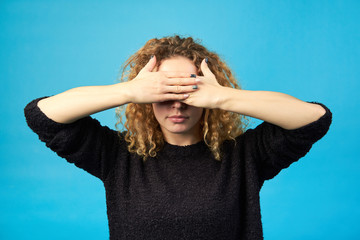 Mood. I don't want to see. Redhead curly girl covering her eyes with hands. Studio shot on blue background.