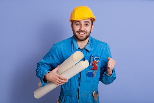 Young handsome bearded man engineer, architect holding rolled up technical drawing, looks happy, celebrating succses of new project, clenching fists, posing isolated over bue studio background.