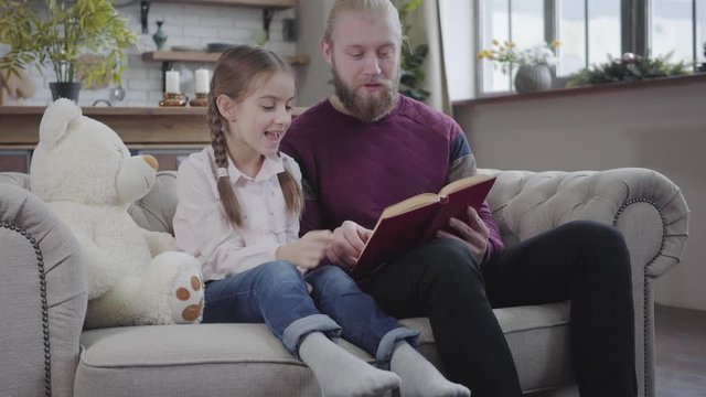 Portrait of intelligent young father reading to his lovely daughter. Interested teen girl listening carefully and showing something in book. Amicable family spending time together at home.