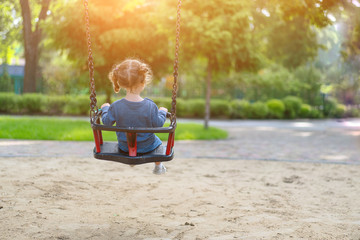Unrecognizable Little Caucasian girl riding swing at playground sunny summer day rear view