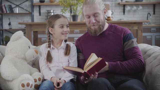 Positive Caucasian father turning page and touching daughter's nose. Cute teenage girl smiling and looking at man. Adult guy educating his child at home. Fun, hobby, lifestyle.