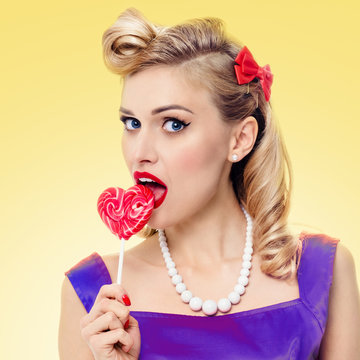 Lovely woman eating heart shape lollipop, pinup style, over yellow background. Caucasian blond girl in retro fashion and vintage concept. Square composition.