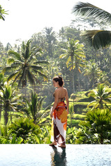 A young girl in an orange bathing suit stands on the edge of the pool against the backdrop of tropical greenery. Bali Indonesia. Ubud.
