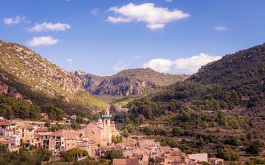 Fototapeta na wymiar Valldemossa church and surrounding town with mountains, forests and blue sky with white clouds, Mallorca, Spain.