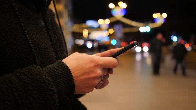 Man order taxi online texting typing smartphone touchscreen display in night city transport stop near road