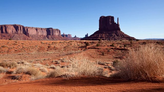 Time Lapse of sunlight and shadows moving across Monument Valley at sunrise.  Camera moves past tumbleweeds with the buttes of Monument Valley in the background.