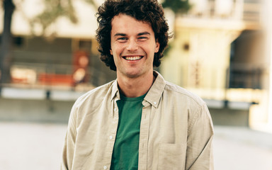 Outdoor horizontal portrait of happy handsome young man smiling and posing for advertisement, looking at the camera, standing at city street. Happy smart student male posing outside. People, emotion