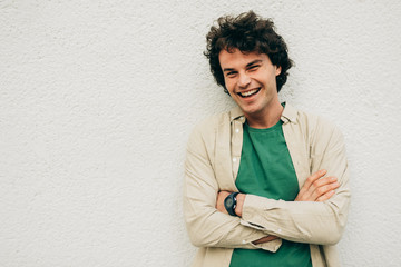 Handsome young man posing for advertisement, smiling and looking at the camera, standing at building concrete wall outdoors. Portrait of happy smart student male with curly hair with crossed arms.