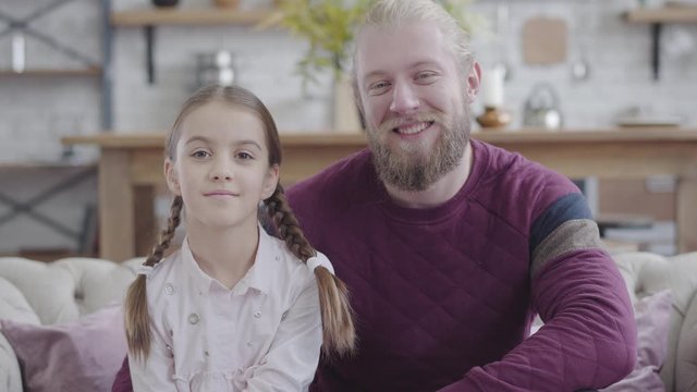 Close-up portrait of happy Caucasian father and daughter looking at camera and smiling. Young bearded man and teenage girl with braided pigtails posing at home. Parenthood, happiness, lifestyle.