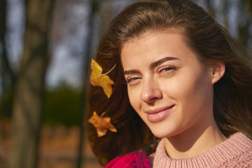 Close-up of female lady, leaves stuck in hair. Portrait of young woman with brown hair, green eyes wearing pink sweater walking in colorful forest or Park. Girl looking into camera has natural beauty.