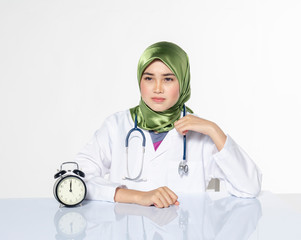 Young doctor with hijab. happy face expression siiting in front laptop ideal for stress management concept.vintage clock, green plant and book on table with reflection