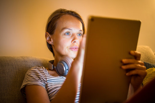 Pretty, young woman working at home in the evening - using her tablet computer as well as bluetooth headphones, writing down some ideas for tomorrow
