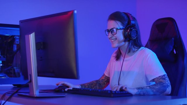 Medium shot of young Caucasian woman with tattoos on her arms wearing headphones and eyeglasses sitting in front of computer screen, playing and talking with her teammates