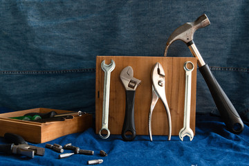 Hand tools such as hammers, pliers, wrenches, screwdrivers, many types of screwdrivers and other tools that are in a wooden box placed on a blue denim background.