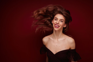 Portrait of pretty positive young long haired lady in black festive top with opened shoulders looking cheerfully at camera and waving her hair, isolated over claret background