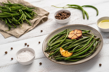 Plate with fried green beans, lemon piece and garlic, uncooked pods on linen napkin, small bowl with olive oil, peppercorns, and sea salt on white wooden table. Healthy food concept