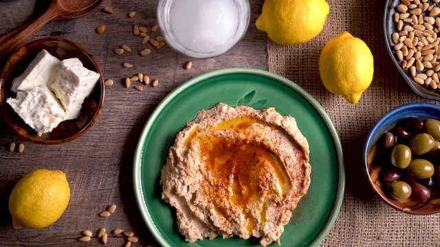 Wide sliding to the right showing a flat lay plate of hummus with feta, olives, glass of ouzo, lemons, pine nuts on a rustic wood table.