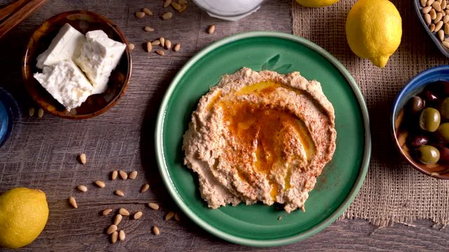 Wide sliding to the right showing a flat lay plate of hummus with feta, olives, glass of ouzo, lemons, pine nuts on a rustic wood table.